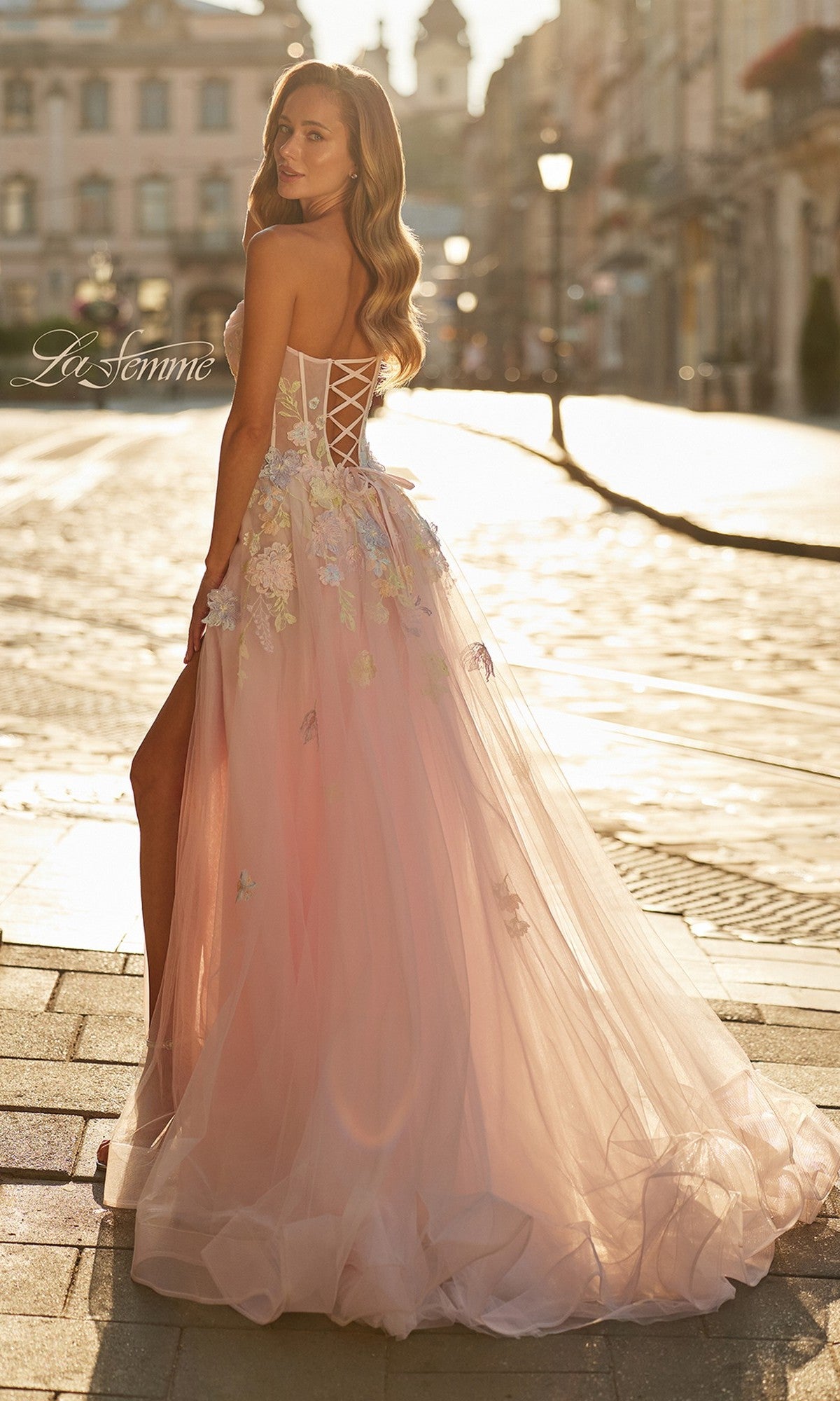 A woman in the La Femme 33432 long prom dress, a strapless light pink ball gown with delicate floral appliqué, stands on a sunlit cobblestone street and looks over her shoulder.